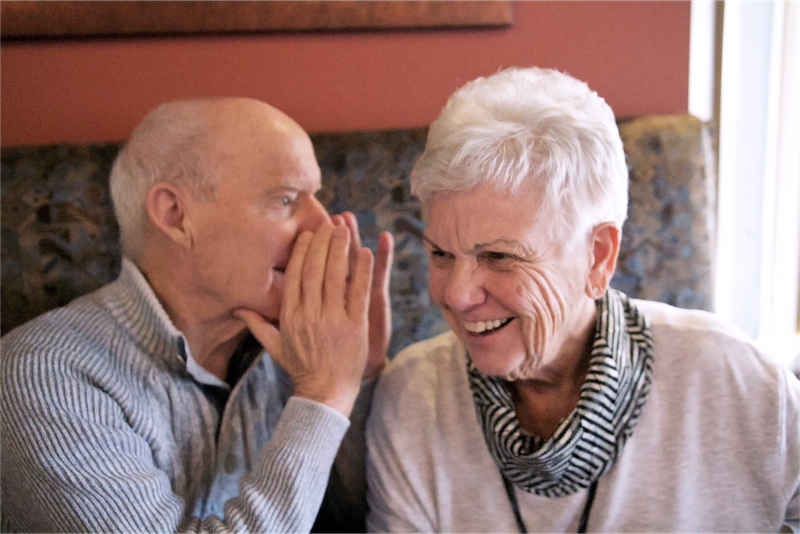 Elderly couple sit side by side laughing while man whispers into woman's ear