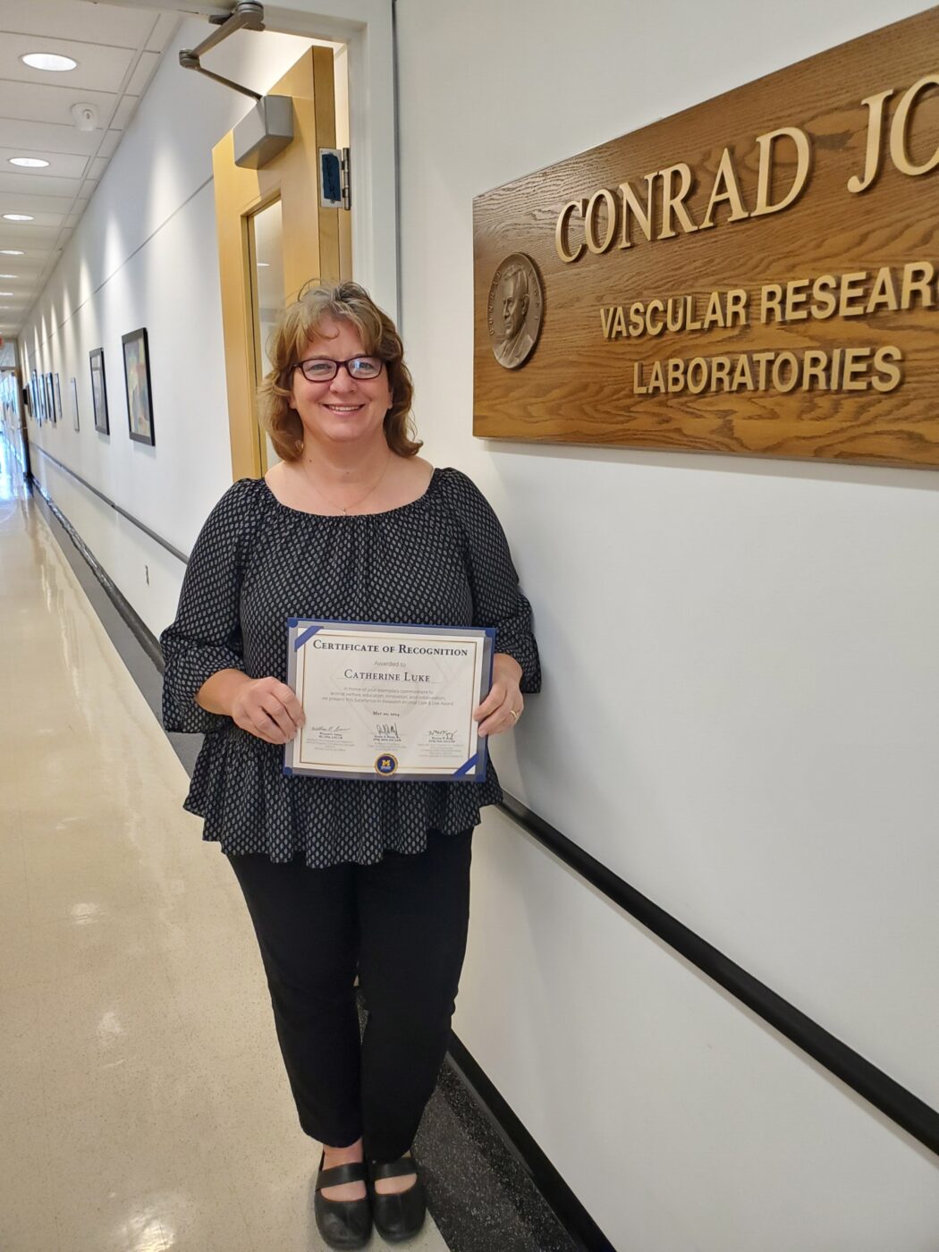 Photo of Cathy Luke, standing in front of the Conrad Jost Lab sign holding a Certificate of Recognition