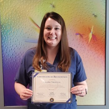 A photo of veterinary technician, Claire Swinton, wearing scrubs in front of a colored background holding a Certificate of Recognition