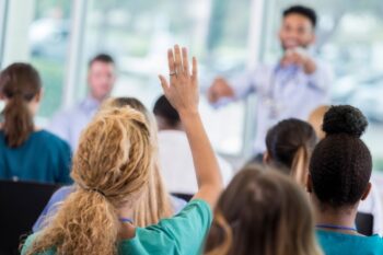 Woman in medical scrubs raises hand to ask a question during a meeting