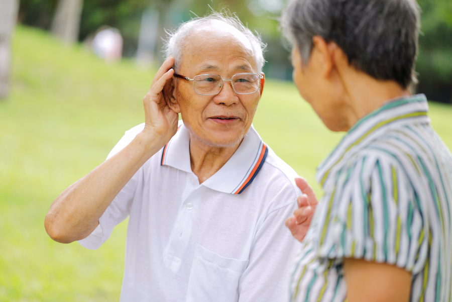 Senior Asian man cups his ear showing difficulty hearing as he listens to his wife during a conversation in the park